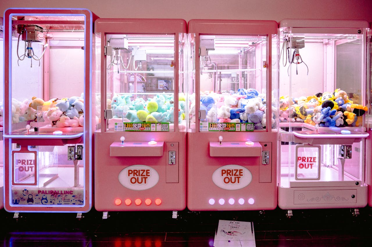 programs-03 Rows of illuminated pink claw machines filled with vibrant soft toys indoors.